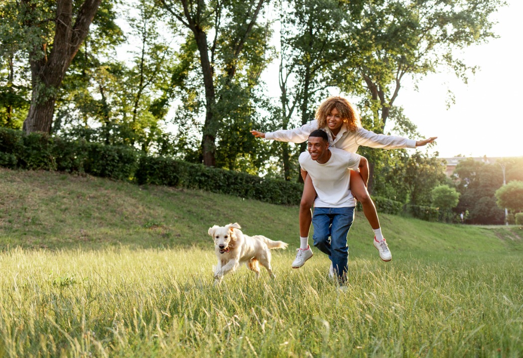 A picture of a women on a mans back run through a field with their dog
