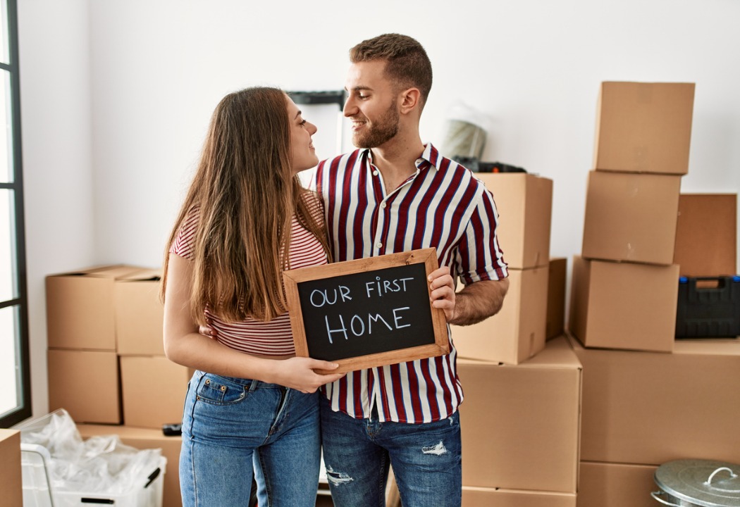 A young couple happily gazing at each other as they hold an "Our First Home" sign, standing among the boxes they have moved into their new home.