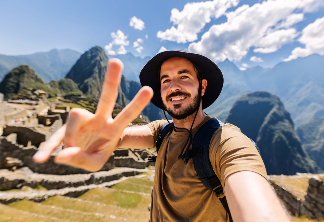 A smiling young man taking a selfie as he gives a peace sign with the back drop of the tourist site he has traveled to.