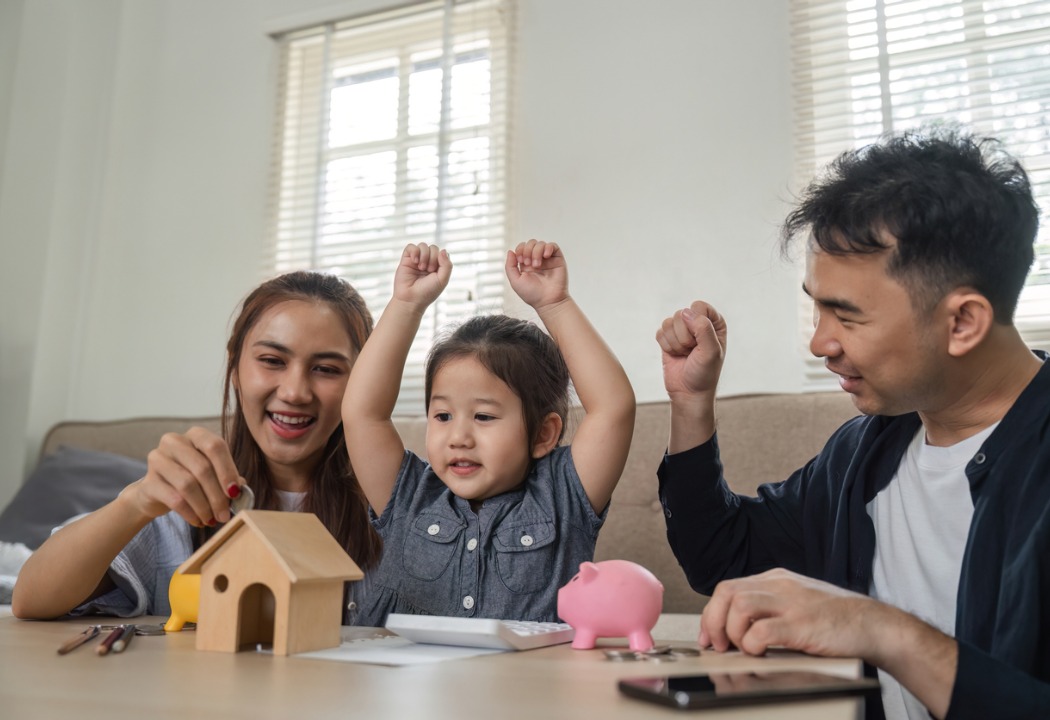 A family of three putting money into a small wooden home shaped piggy bank