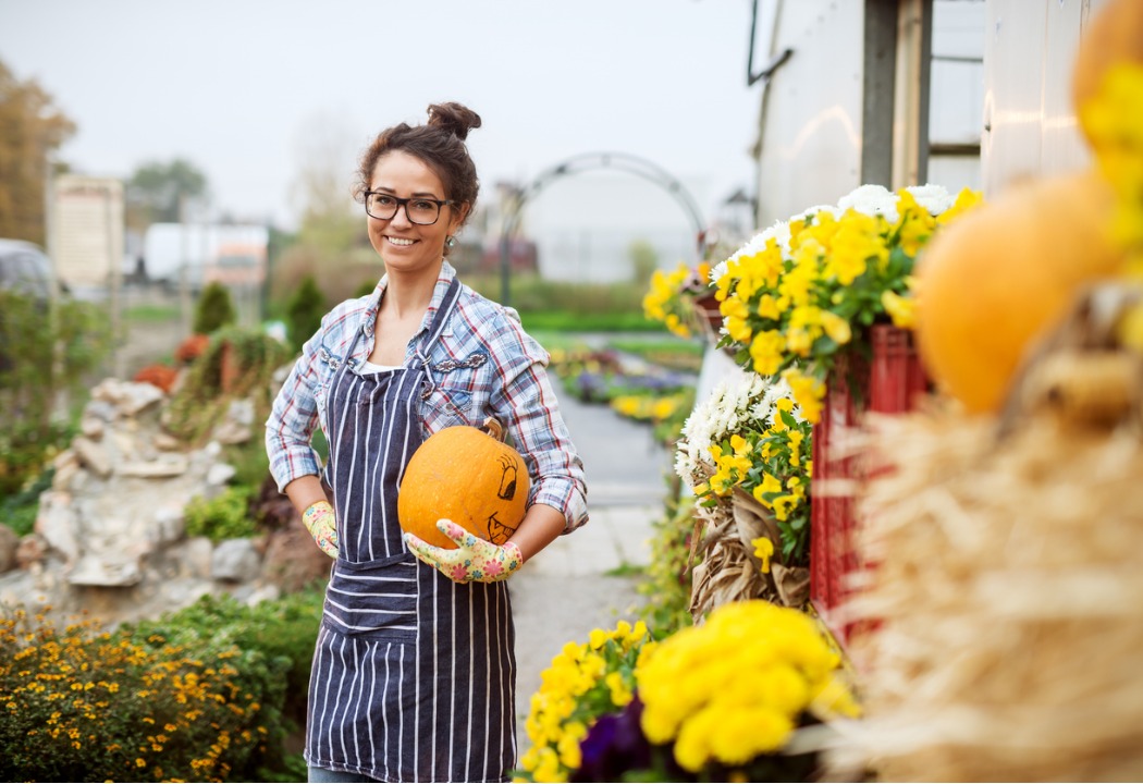 A woman standing next to a greenhouse holding a pumpkin and smiling 