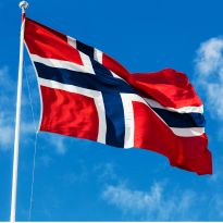 The red Norwegian Flag featuring a blue Nordic cross outlined in white blowing in the wind suspended from a silver pole on partially cloudy day.